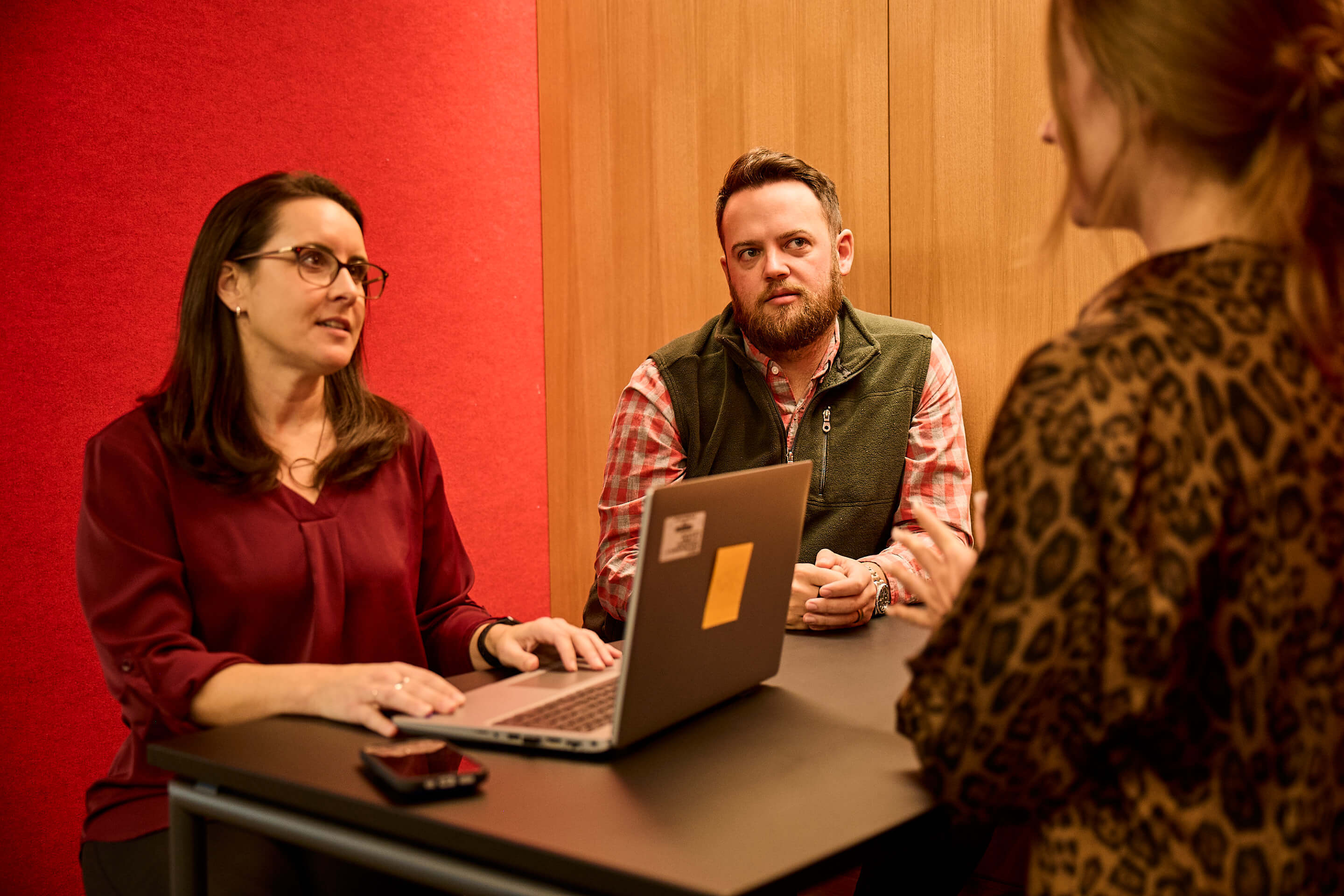 Three colleagues sitting together at a table, engaged in discussion while using a laptop.