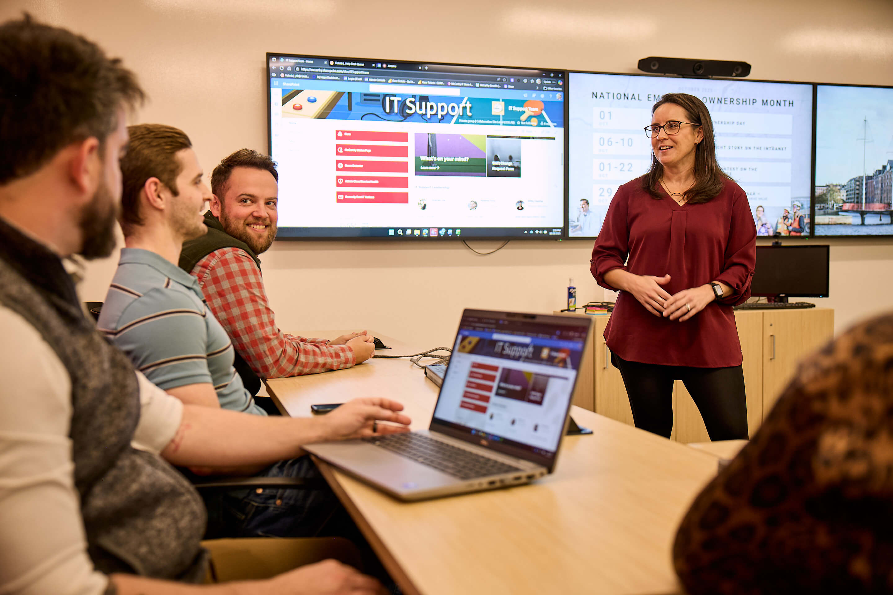 A woman addresses a group of people while standing in front of three screens.