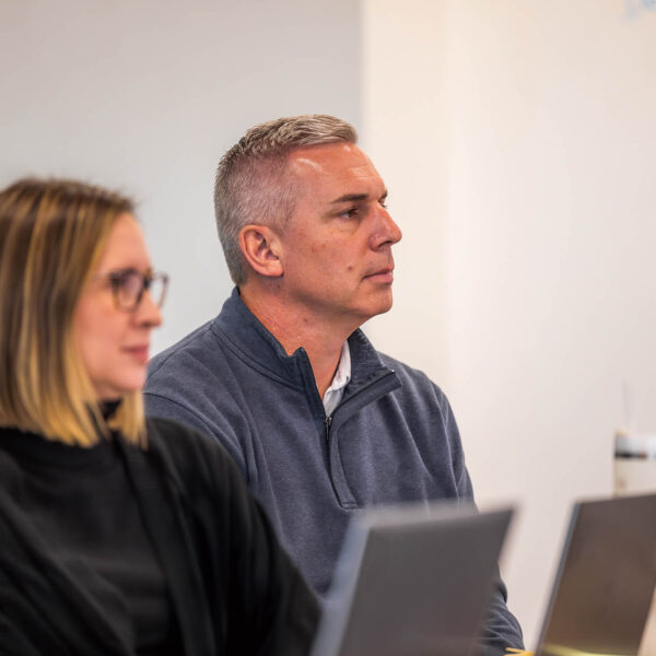 A man in a blue shirt sitting in front of a laptop looking at a presenter in a meeting.