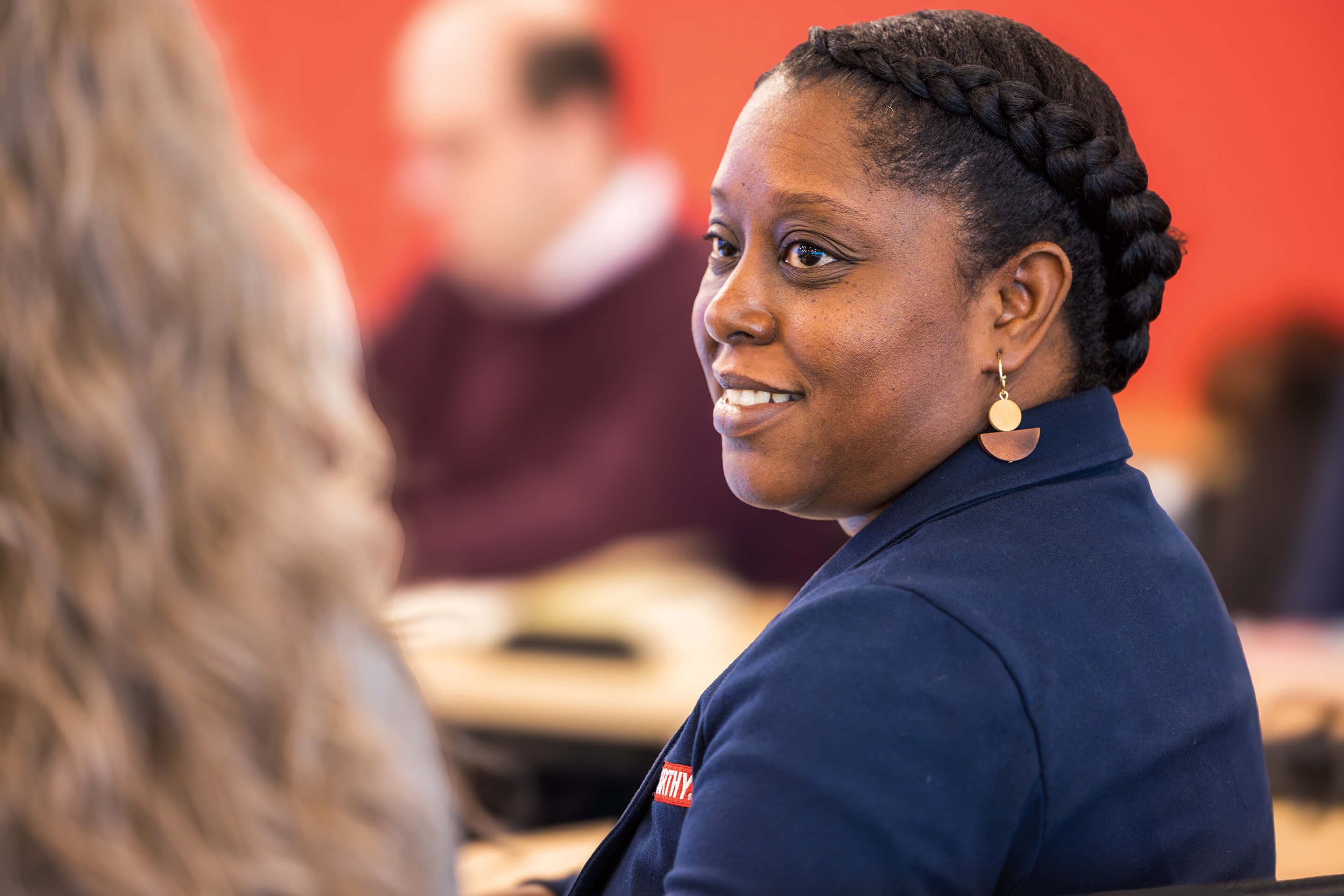 A woman in a blue jacket is seated at a table, appearing focused and thoughtful.